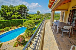 A poolside area with a view of a lush landscape, a covered patio with a table and chairs, and a railing along the pool.