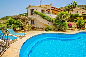 A poolside view of a Mediterranean-style villa with a curved swimming pool, a staircase leading to a balcony, and lush landscaping.
