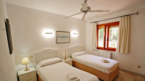 A bedroom with two beds, a ceiling fan, a window with curtains, a nightstand with a lamp, and a framed picture on the wall. - Nessa - well furnished villa with panoramic views in Benitachell (Benitachell)