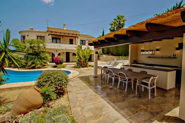 A poolside area with a dining setup, a pergola, and a view of a multi-story building.