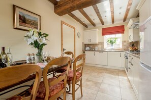 A dining area with wooden chairs and a table set for two, a kitchen with white cabinets and a red window valance, and a painting on the wall.