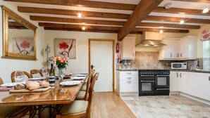 Un coin repas avec une table en bois dressée pour le repas, une cuisine avec des meubles blancs et des appareils électroménagers, et un plafond avec des poutres apparentes.