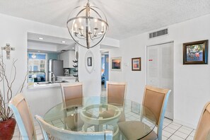 A dining area with a glass table and chairs, a chandelier, and a view into the kitchen.