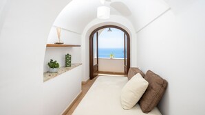 Une pièce étroite avec un plafond voûté, un petit coin salon avec des coussins, une étagère ornée d’objets décoratifs et une vue sur la mer par une porte en arc de cercle.