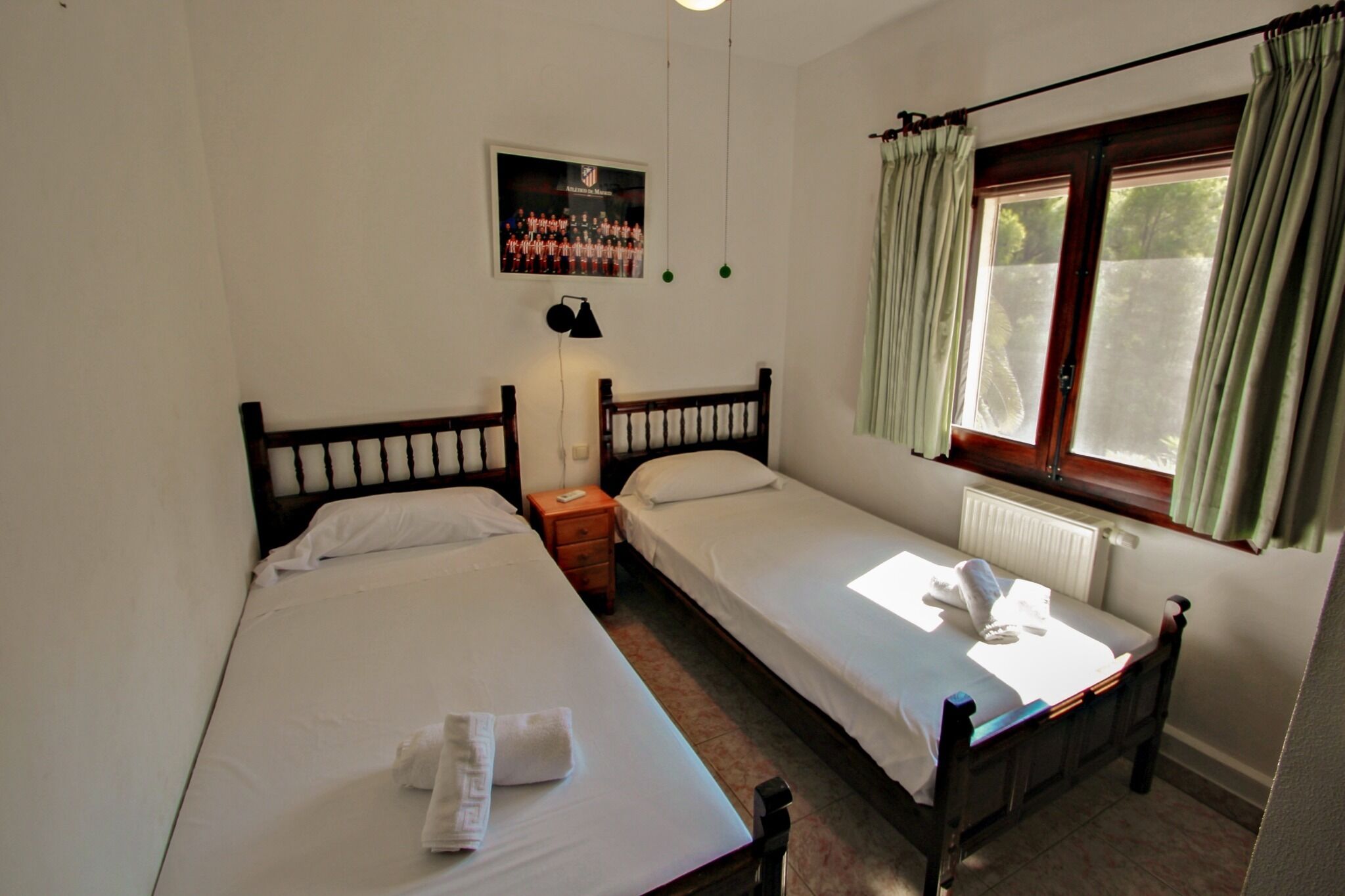 Une chambre avec deux lits simples, une table de chevet, une lampe, une fenêtre avec des rideaux et une affiche au mur.