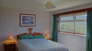A bedroom with a bed, two bedside lamps, a framed picture on the wall, and a window with curtains. - Steading Holidays - Ceol na Mara (Acharacle)