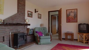 A living room with a brick fireplace, a green armchair, a television, and a bookshelf. - Steading Holidays - Ceol na Mara (Acharacle)