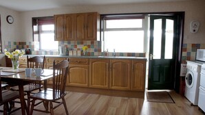 A kitchen with wooden cabinets, a dining table with chairs, a washing machine, and a window with a view. - Steading Holidays - Ceol na Mara (Acharacle)