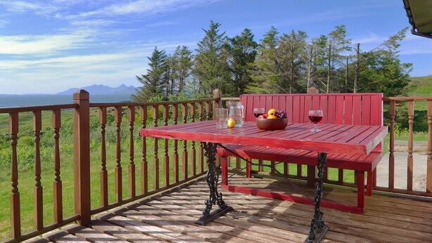A red wooden table with a bowl of fruit and two wine glasses on a deck with a view of mountains and trees. - Steading Holidays - Ceol na Mara (Acharacle)