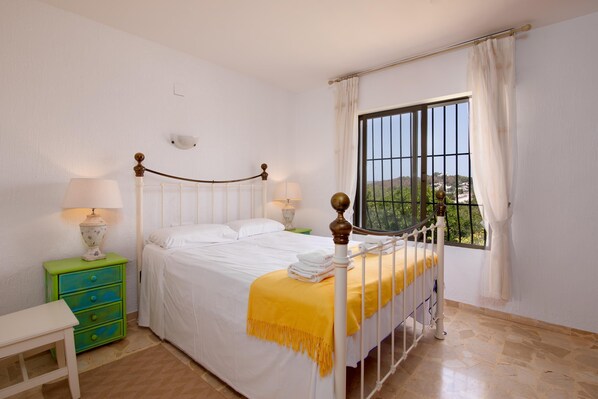 A bedroom with a bed, bedside lamps, a green nightstand, a window with curtains, and a view of greenery outside. - El Tesorito Javea - Javea, Costa Blanca (Javea)