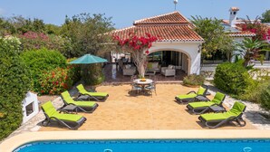A pool area with lounge chairs and a table, surrounded by greenery and a building with a tiled roof. - El Tesorito Javea - Javea, Costa Blanca (Javea)