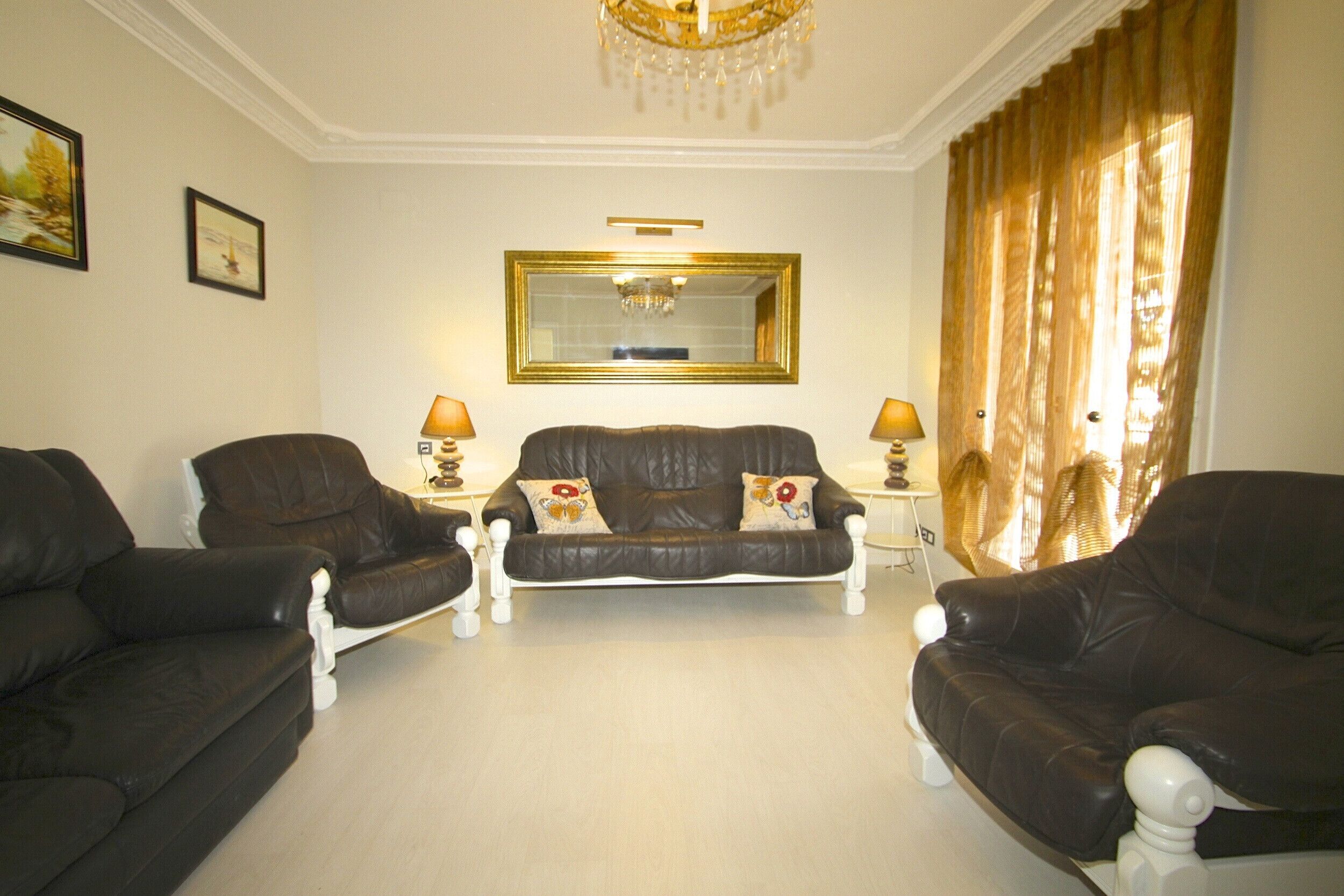 A living room with a brown leather sofa set, two side chairs, a small table with a lamp, a chandelier, and a large mirror on the wall.