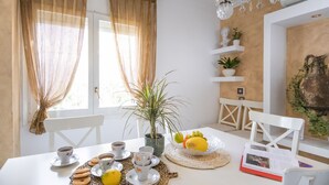 Een ontbijtruimte met een gedekte tafel, inclusief gebak, koffie en fruit. Er zijn witte stoelen, een kroonluchter en decoratieve planten.