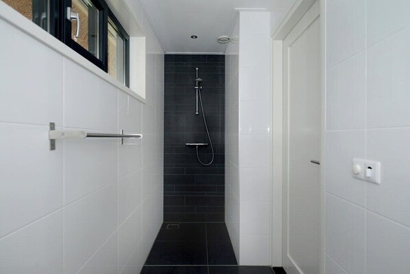 A narrow, tiled bathroom with a shower, white walls, and a door with a handle.