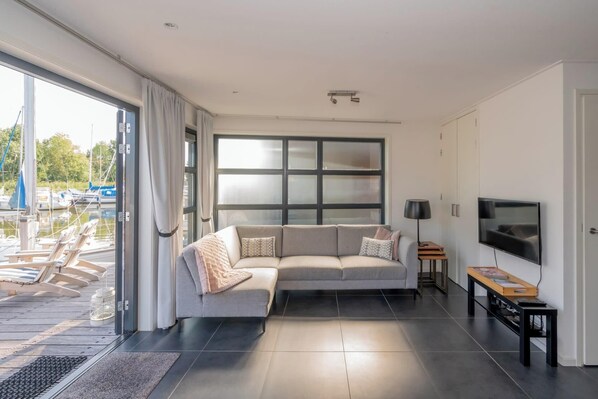 A modern living room with a large window, a sofa, a TV, and a small table. - Enjoy a wonderful stay in this stylish waterfront villa for 6 people, situated in a unique location (De Heen)
