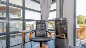 Una habitación moderna con ventanas amplias, un sillón de cuero, una mesita redonda y una estufa de leña.