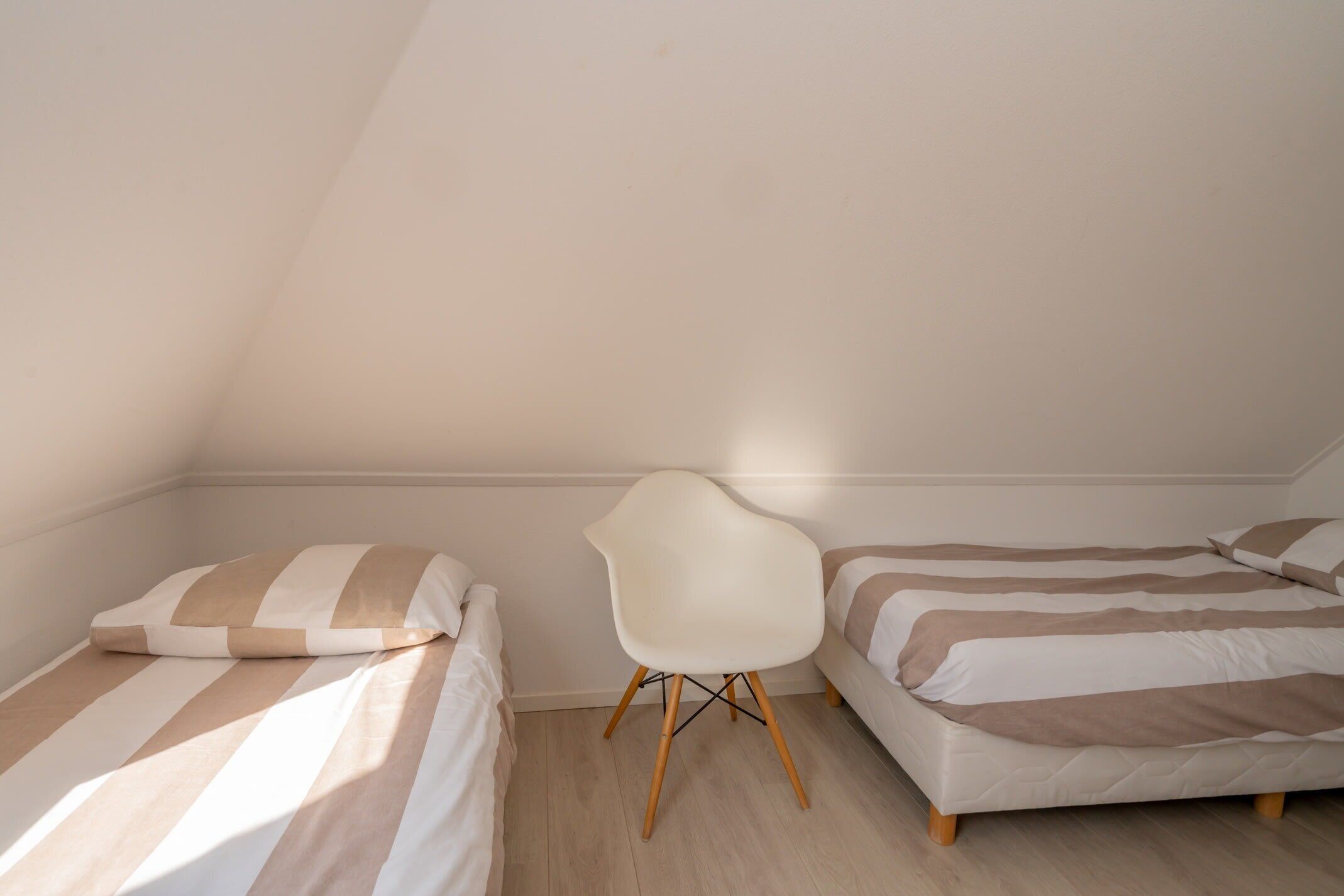 A small, well-lit room with two beds, a white chair, and a sloped ceiling.