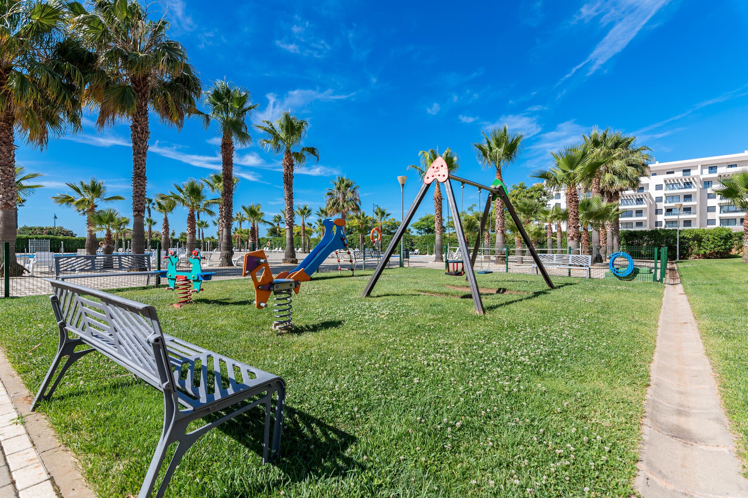 A playground with swings and slides, surrounded by palm trees and a bench.