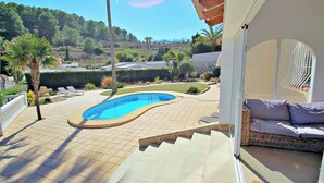 A pool area with a curved pool, a seating area with cushions, and a view of a hilly landscape. - Alldo - modern hillside villa with pool & views (Moraira)