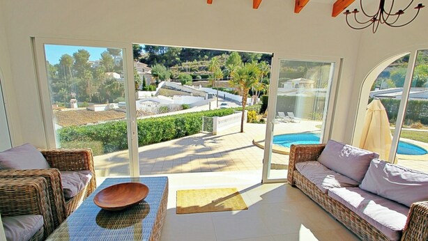 A sunlit room with wicker furniture, a glass door leading to a pool, and a view of a residential area. - Alldo - modern hillside villa with pool & views (Moraira)
