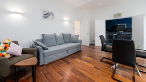 A modern living room with a grey sofa, a small table, and a black chair. There is a kitchen area visible through an open door.
