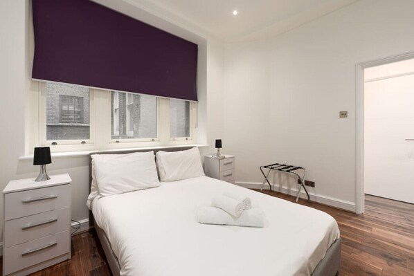 Ein Schlafzimmer mit einem Bett, zwei Nachttischen, einem Fenster mit Jalousien, einem Heizkörper und einer Tür.