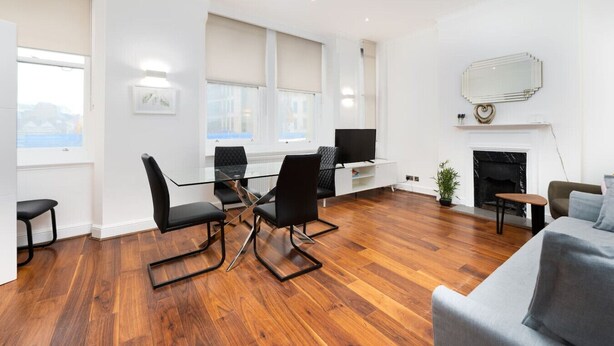 A modern living room with a glass dining table, black chairs, a grey sofa, a TV, and a fireplace.