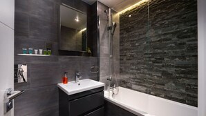 A modern bathroom with a glass-enclosed shower, a white sink with a mirror above, and dark tiled walls.