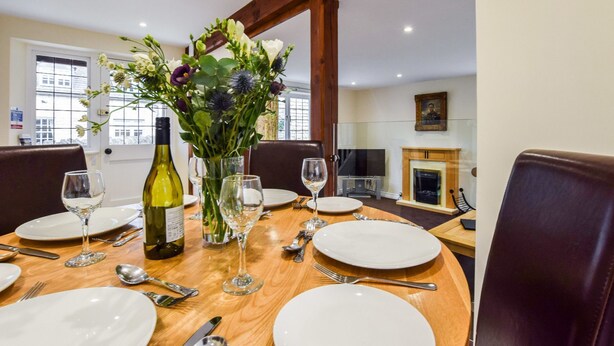 A dining table set for four with white plates, silverware, and wine glasses. A bottle of wine and a vase with flowers are on the table. The room has wooden chairs, a fireplace, and a large window. - A sudeley castle cottage that sleeps 4 guests  in 2 bedrooms (Cheltenham)