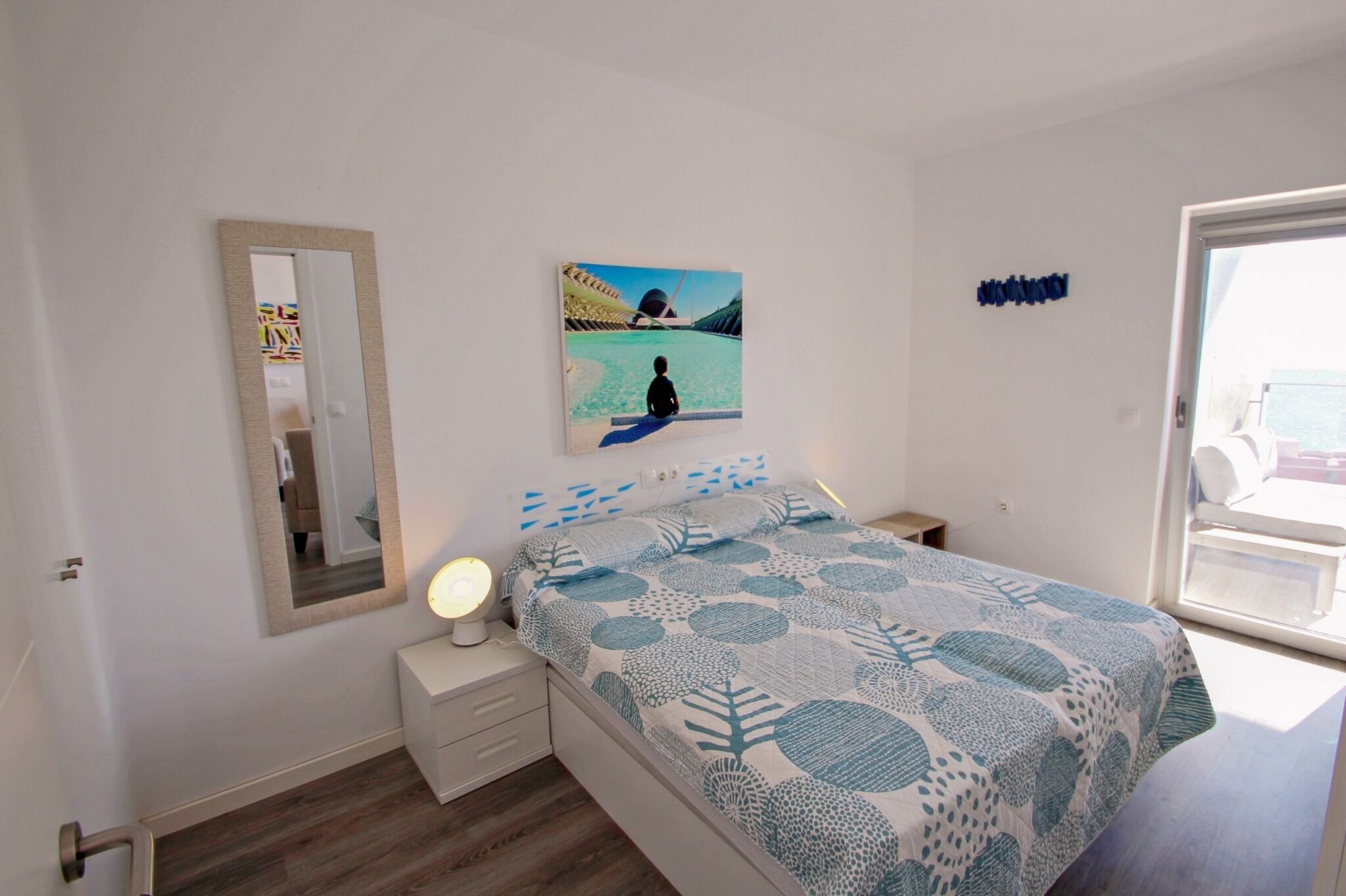 A bedroom with a bed, a bedside table with a lamp, a mirror, a painting on the wall, and a window.