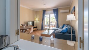 Una habitación de hotel moderna con zona de comedor, sofá, televisor y ventana con cortinas.