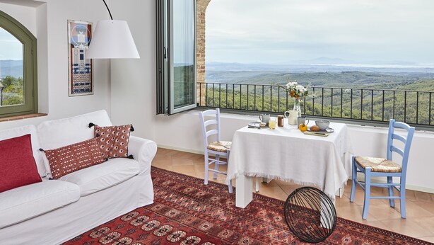Une pièce avec un canapé blanc, un coussin rouge, un tapis à motifs, une table à manger avec des chaises, et une vue sur un paysage montagneux depuis un balcon.