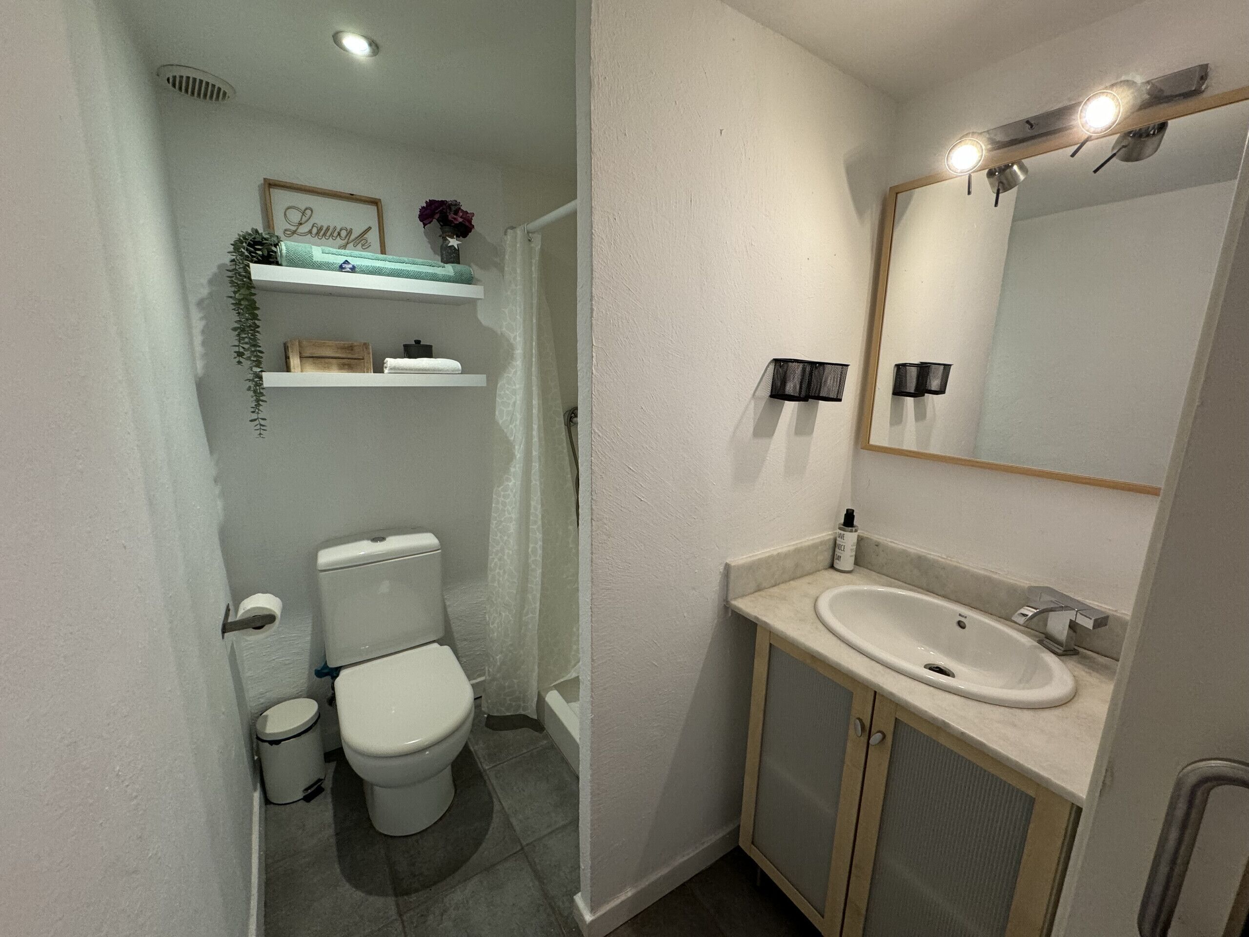 A bathroom with a toilet, sink, mirror, and shelves with toiletries.