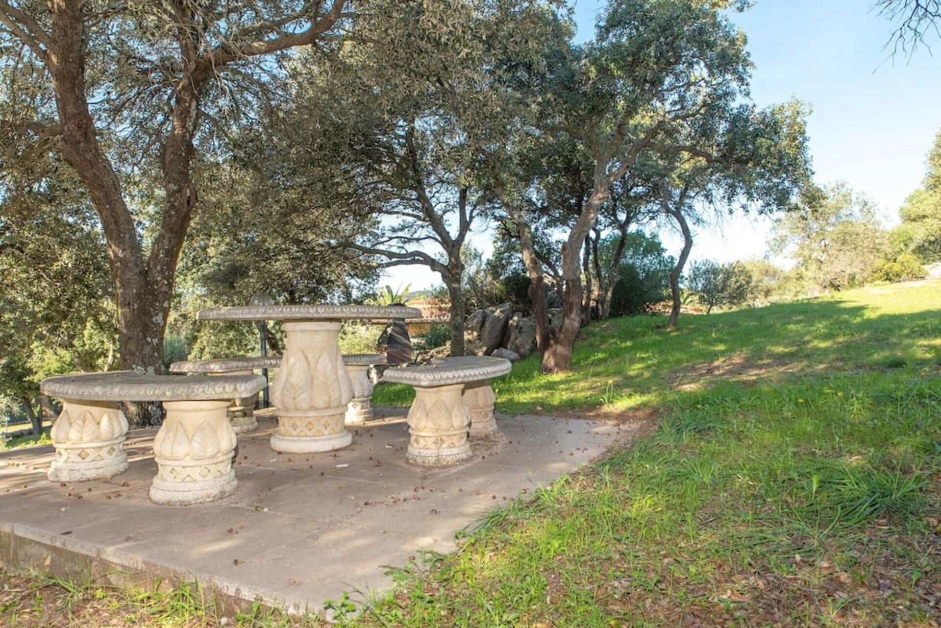A stone outdoor seating area with a table and chairs, surrounded by trees and grass.