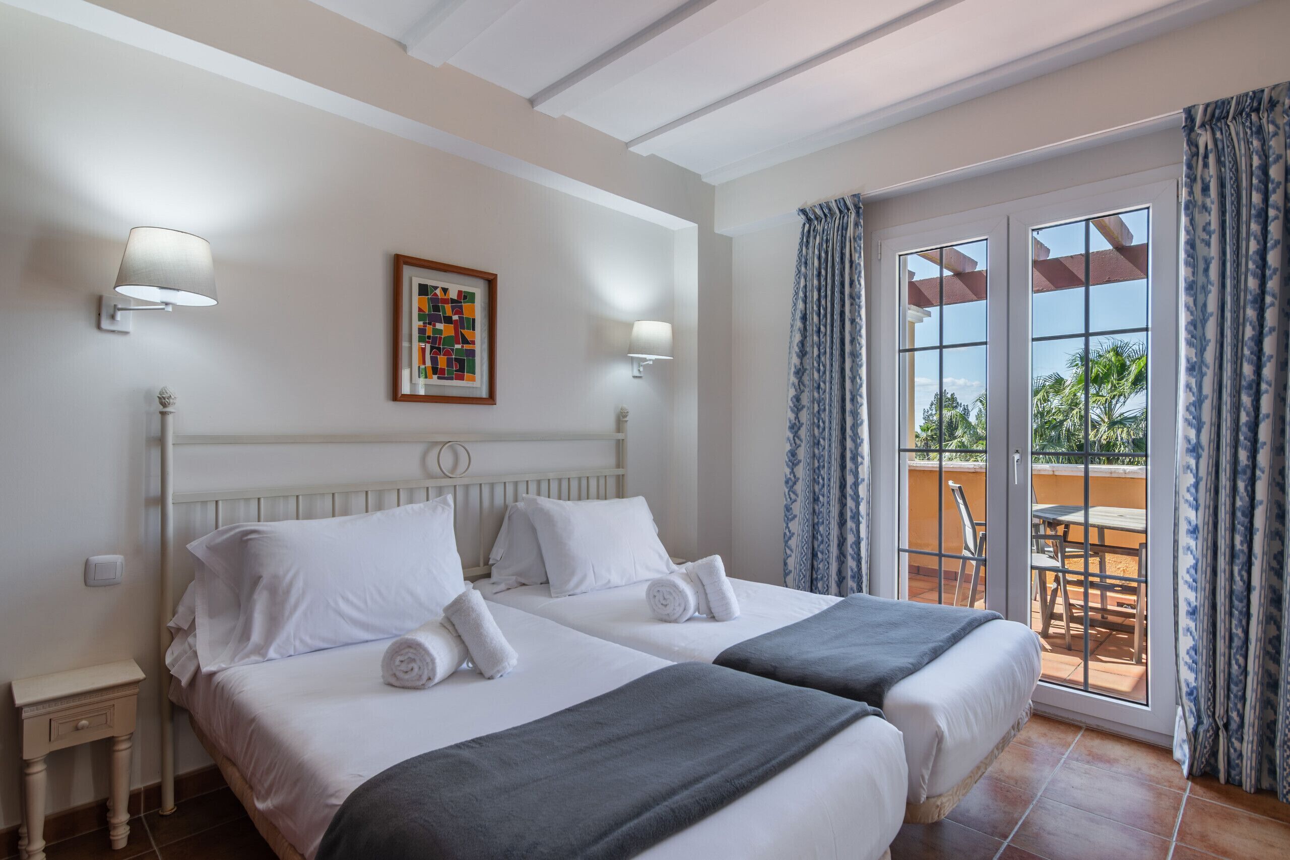 A hotel room with two beds, a balcony with a table and chairs, and a view of palm trees.