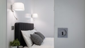 Une chambre moderne avec une tête de lit noire, des coussins blancs et gris, une petite plante en pot et deux lampes fixées au mur.