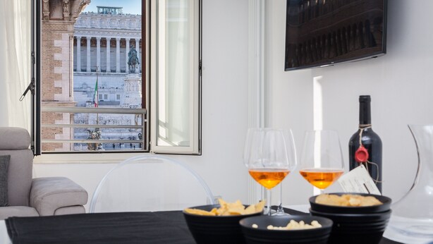 Un coin repas avec une table dressée pour deux personnes, deux verres de vin, une bouteille de vin et des bols remplis de collations. Une fenêtre offre une vue sur un bâtiment historique orné de colonnes et de statues.