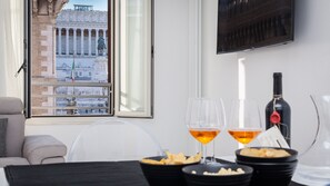 Un coin repas avec une table dressée pour deux personnes, deux verres de vin, une bouteille de vin et des bols remplis de collations. Une fenêtre offre une vue sur un bâtiment historique orné de colonnes et de statues.
