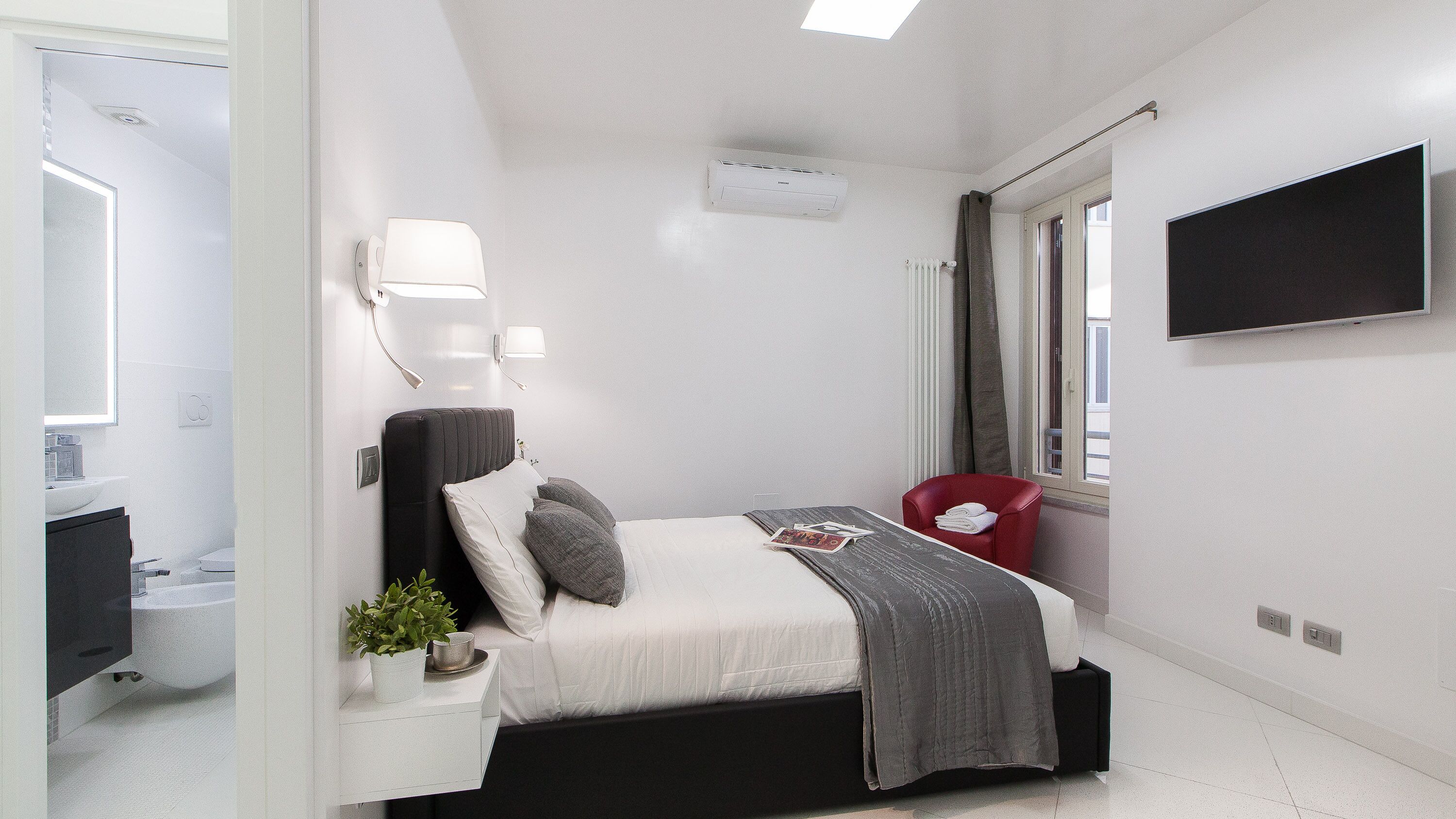 A modern hotel room with a bed, bedside table, lamp, and a TV mounted on the wall.