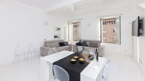 Un salon moderne avec un coin repas, comprenant une table blanche dressée avec des verres et des amuse-gueules, deux canapés avec des coussins et une grande fenêtre donnant sur un immeuble.