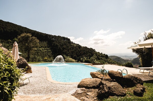 A swimming pool with a fountain, surrounded by rocks and greenery, with a view of a forested hill in the background.