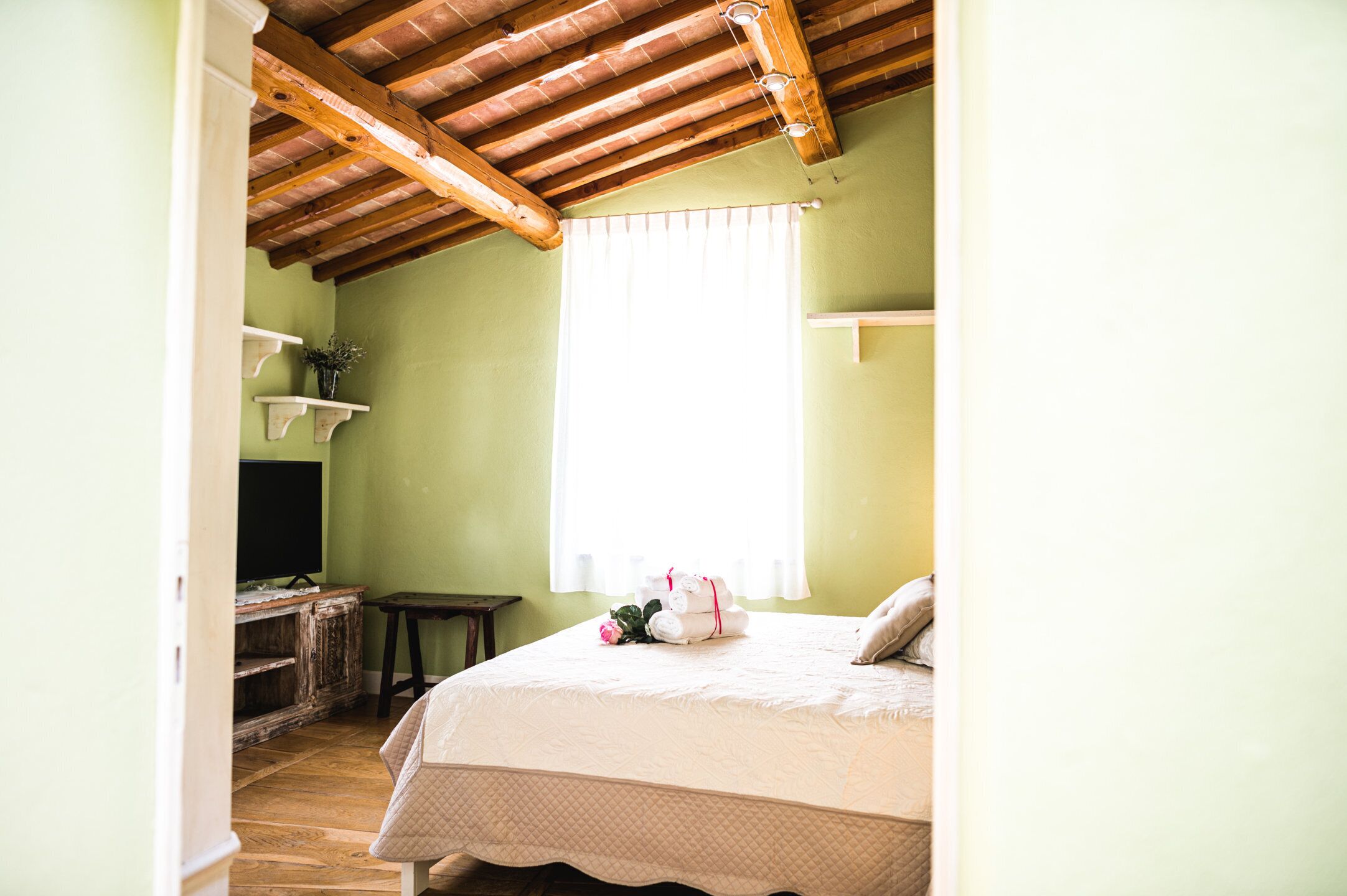 Un dormitorio con techo de madera, una cama con cubrecama blanco, una mesita de noche, una planta pequeña en maceta y una ventana con cortinas blancas.
