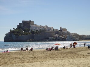 Ein Strand mit Menschen, die unter Sonnenschirmen entspannen, ein historisches Gebäude im Hintergrund und ein klarer, blauer Himmel.