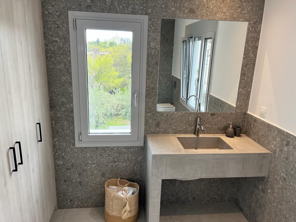 A bathroom with a concrete sink, a large mirror, and a window with a view of greenery.