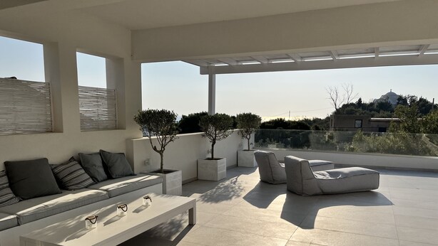A modern outdoor patio with a sofa, chairs, and a table. There are potted plants and a view of the surrounding landscape.