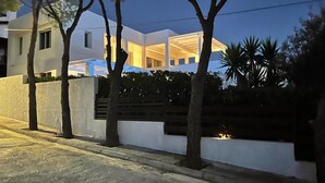 A modern house with a white exterior, a visible interior with large windows, and a well-maintained garden with trees and shrubs.