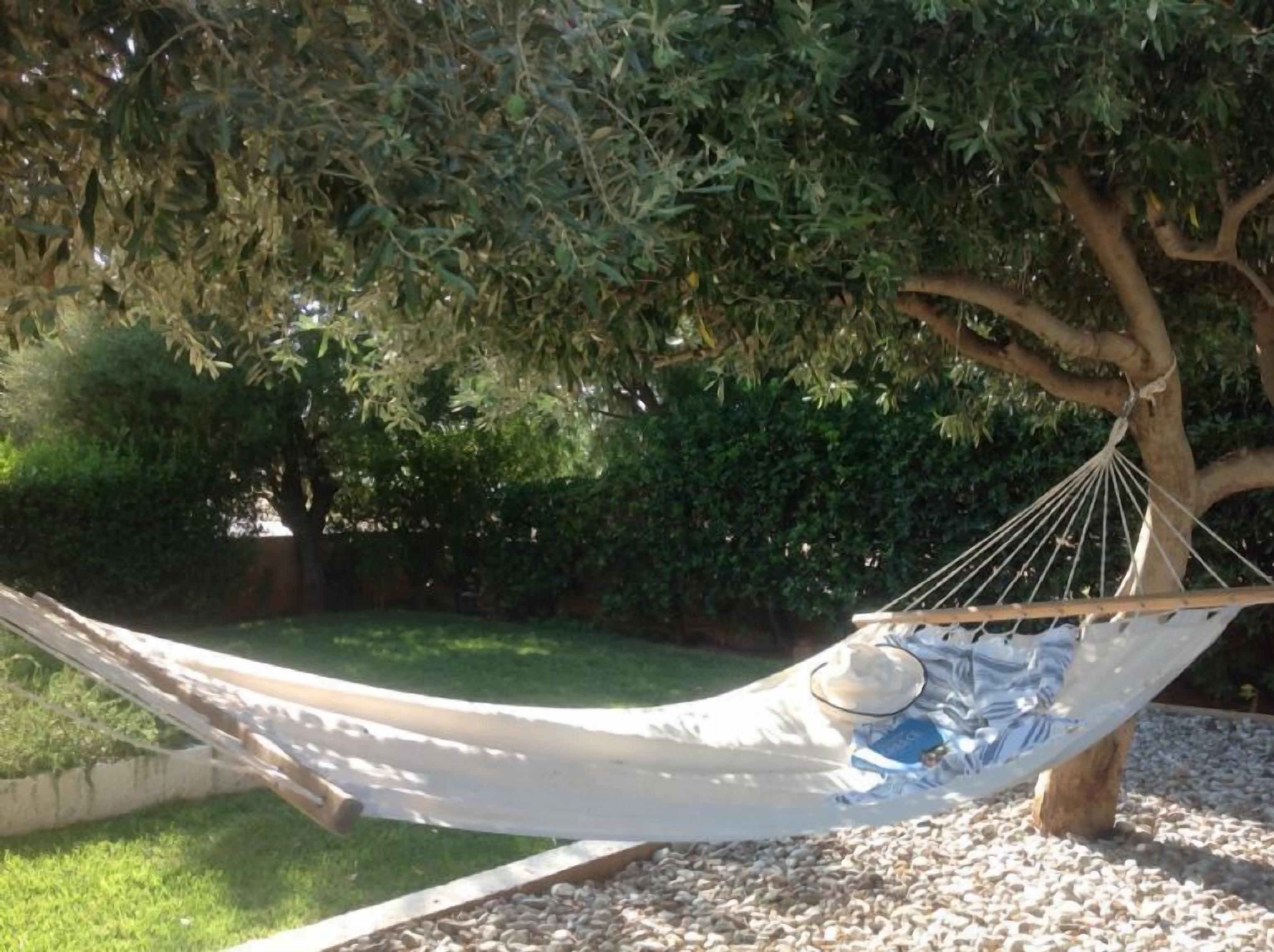 A hammock under a tree in a garden with pebbles and grass.