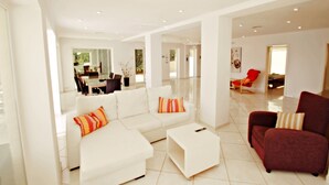 Un salón espacioso con un sofá blanco, un sillón rojo y una mesa de centro. En el fondo, se observa un comedor con una mesa y sillas.