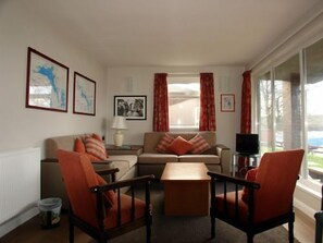 Una sala de estar con un sofá, dos sillones, una mesita de centro y un televisor. Hay cuadros enmarcados en la pared, una lámpara sobre una mesita auxiliar y una ventana con cortinas.