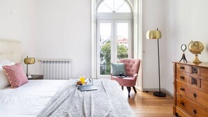 A bedroom with a bed, a dresser, a chair, a radiator, and a window with a view of buildings.